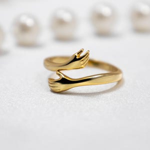 May include: A gold ring with two hands clasped together, forming a circle. The ring is on a white background.