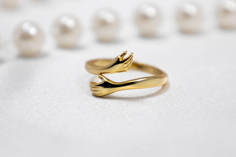 May include: A gold ring with two hands clasped together, forming a circle. The ring is on a white background.