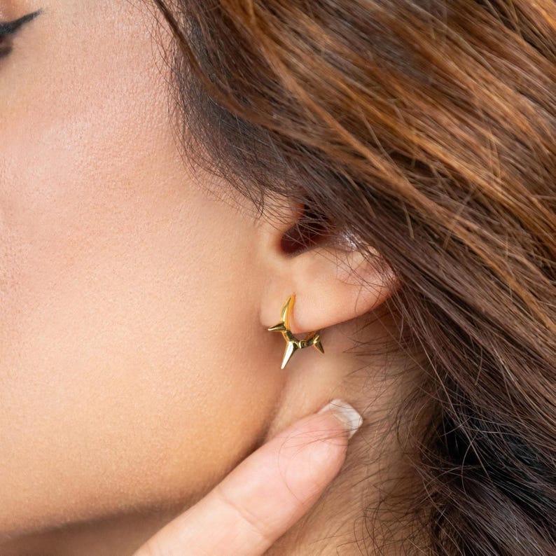 May include: Gold hoop earrings with a pointed star design. The earrings are worn in the ear of a person with brown hair.