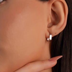 May include: Silver hoop earrings with a rectangular shape. The earrings are worn in the earlobes of a person with dark hair.