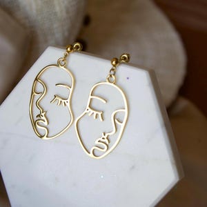 May include: Gold-colored earrings featuring a minimalist design of two faces with closed eyes. The earrings are suspended from small, round studs. The faces are outlined with delicate lines, creating a modern and artistic aesthetic.