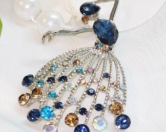 May include: A silver-toned brooch in the shape of a ballerina. The dancer's body is adorned with blue and brown gemstones, with a skirt of dangling crystals. The brooch is set against a white background with pearl accents.