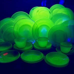 May include: A collection of vintage glassware including multiple plates, saucers, and cups, all glowing with a bright neon green fluorescence under ultraviolet light. The pieces feature a ribbed, scalloped edge design and a translucent appearance.