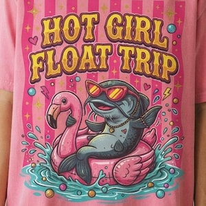 May include: A pink short-sleeved t-shirt featuring a graphic print with the text "HOT GIRL FLOAT TRIP" in yellow letters. The design shows a cartoon catfish wearing sunglasses and a necklace, floating in a pink flamingo pool float in water.
