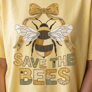 May include: Yellow t-shirt with a bee graphic and the text "SAVE THE BEES." The bee design includes a bow and patchwork patterns in brown, black, and white. The text and bow have a patchwork design in yellow and green.