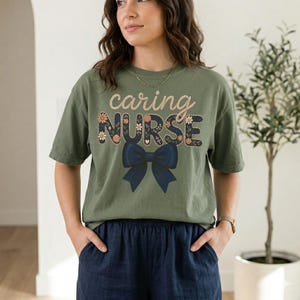May include: Olive green t-shirt featuring the text "caring NURSE" in a floral design and a navy blue bow. A casual style t-shirt suitable for everyday wear.