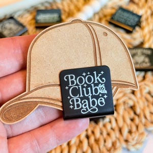 May include: A wooden baseball cap-shaped bookmark with a black rectangular clip. The clip has the text "Book Club Babe" in white script. The bookmark is light brown and appears to be laser cut.