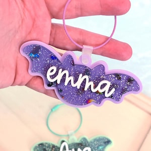 May include: A purple bat-shaped keychain with the name "emma" in white script. The keychain has a glittery interior and a pink wire loop. A similar green keychain with the name "Ava" is also visible.
