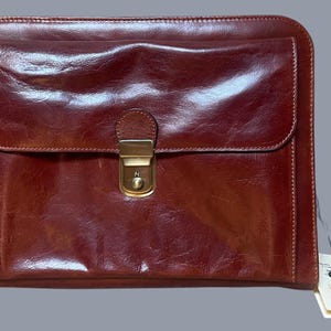 May include: A rectangular dark reddish-brown leather document case featuring a smooth, polished finish and visible stitching along the edges. The front flap is secured with a central brass-toned metal push-lock clasp. A white paper tag is attached.