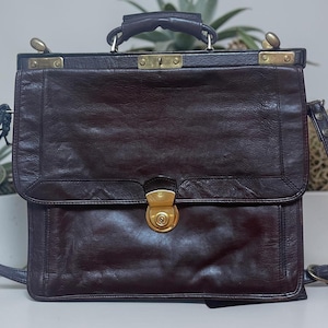 Puede incluir: Un maletín de cuero marrón oscuro con una forma rectangular estructurada, un asa superior y una correa de hombro ajustable. El bolso incluye una solapa frontal asegurada por un cierre de presión de metal dorado y herrajes a juego.