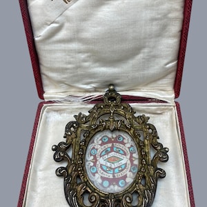 Pode incluir: Uma antiga relíquia religiosa numa caixa de veludo vermelho. A relíquia está numa moldura dourada ornamentada com uma imagem central e texto. A caixa tem um forro de cetim branco e uma etiqueta que diz "A. Peroni VERONA".