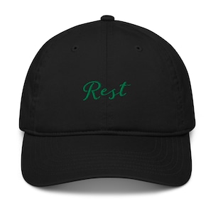 Op de afbeelding: Zwarte baseballcap met een gebogen klep. Het woord "Rest" is in groen script op de voorkant geborduurd. De cap is gemaakt van een zachte, ademende stof.