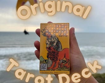 May include: A hand holds a yellow tarot card with a woman and a lion. The card is from the "Tarot Original 1909" deck by Arthur Edward Waite and Pamela Colman Smith. The text "Original" is written in large, yellow letters above the card.
