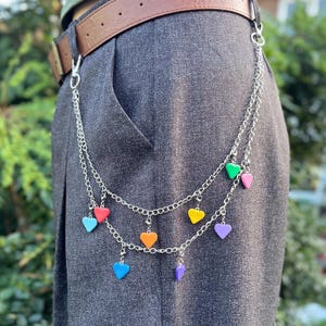 May include: A silver chain belt with colorful heart charms. The belt is attached to a brown leather belt and hangs down over gray pants. The hearts are red, orange, yellow, green, blue, and purple.
