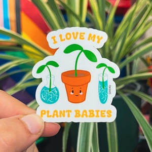 May include: A white sticker with the text "I LOVE MY PLANT BABIES" in orange. The sticker features three illustrations of plants: a potted plant with a smiling face and two plants in glass containers with blue liquid.