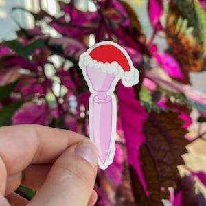 May include: A pink sticker featuring a cartoon design of a pink object with a red Santa hat and white trim. The sticker is held in a hand against a background of green and pink leaves.