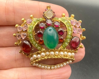 May include: A gold crown brooch with a green stone in the center and red, pink, and green stones surrounding it. The crown is decorated with pearls.
