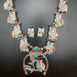 Puede incluir: Un collar de plata con un gran colgante de búho y pendientes de búho más pequeños. El collar y los pendientes están decorados con incrustaciones de turquesa, coral y concha. El colgante de búho tiene forma de herradura.