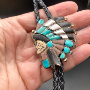 May include: A bolo tie featuring a central inlay pendant shaped like a profile of a person wearing a feathered headdress. The inlay consists of turquoise, black stone, white shell, and pink stone segments set in silver, attached to a braided black cord.