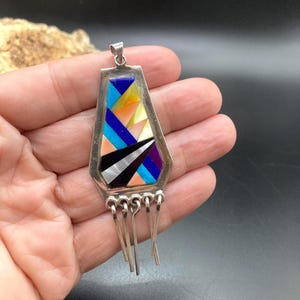 Op de afbeelding: Een zilveren hanger met een geometrische inleg van veelkleurige steen- en schelpsegmenten in tinten blauw, zwart, wit, oranje en paars. De hanger heeft een trapeziumvorm met vijf bungelende zilveren metalen staafjes aan de onderkant.