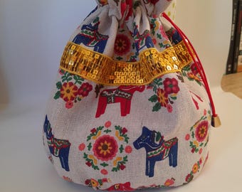 Pode incluir: Uma bolsa de cordão branco com um padrão floral vermelho, azul e amarelo que apresenta um desenho repetido de um cavalo Dala sueco. A bolsa tem uma guarnição de lantejoulas douradas.
