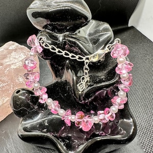 May include: A pink and white beaded bracelet with a silver clasp. The bracelet is made of small, irregular shaped pink beads and white pearl beads.