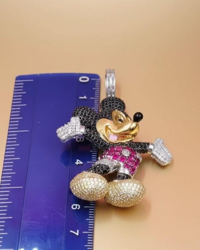 Puede incluir: Un colgante con un dise&ntilde;o estilizado de Mickey Mouse. El colgante tiene una cara dorada, un cuerpo negro y detalles en rojo y blanco. El colgante mide aproximadamente 5 cm de altura.