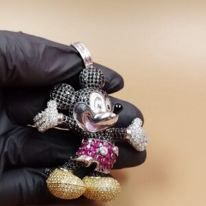 Puede incluir: Un colgante de Mickey Mouse con cara plateada, cuerpo negro y blanco y zapatos dorados. Los pantalones cortos son de color rosa vibrante y las manos est&aacute;n incrustadas con piedras preciosas blancas. El colgante lo sostiene una mano enguantada de negro.
