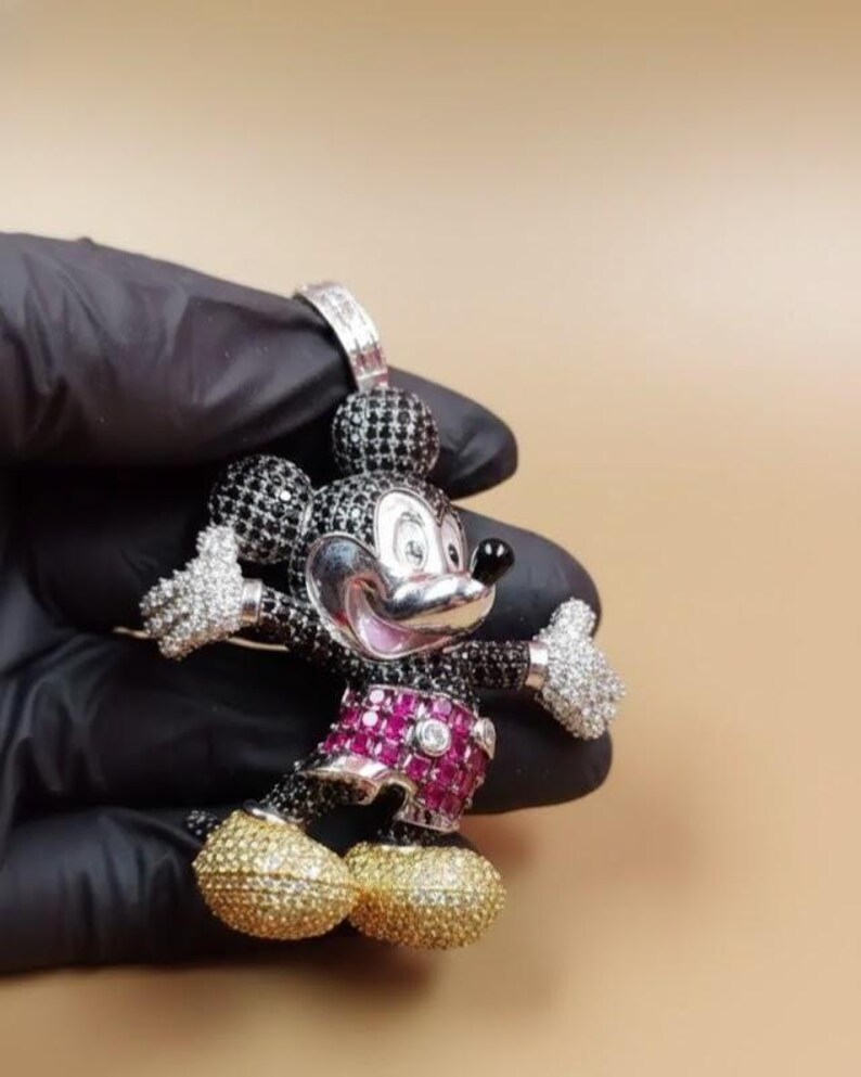 Puede incluir: Un colgante de Mickey Mouse con cara plateada, cuerpo negro y blanco y zapatos dorados. Los pantalones cortos son de color rosa vibrante y las manos est&aacute;n incrustadas con piedras preciosas blancas. El colgante lo sostiene una mano enguantada de negro.