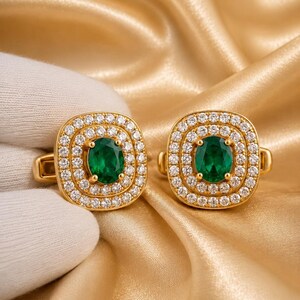 May include: A pair of gold cufflinks featuring a central oval-cut green gemstone surrounded by two concentric halos of small, clear, round-cut white stones. The square-shaped settings have rounded corners and a polished gold-tone metal finish.
