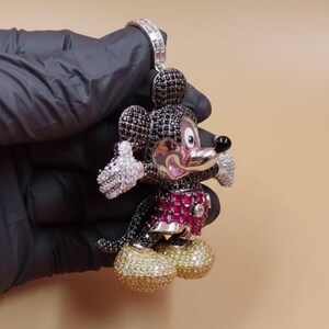Puede incluir: Un colgante con dise&ntilde;o de Mickey Mouse. El colgante est&aacute; incrustado con piedras preciosas negras, blancas, rosas y doradas. La figura lleva un atuendo rojo y rosa y zapatos dorados. El colgante se sujeta con una fianza plateada.