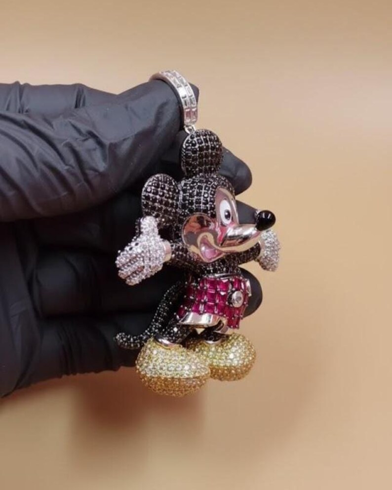 Puede incluir: Un colgante con dise&ntilde;o de Mickey Mouse. El colgante est&aacute; incrustado con piedras preciosas negras, blancas, rosas y doradas. La figura lleva un atuendo rojo y rosa y zapatos dorados. El colgante se sujeta con una fianza plateada.