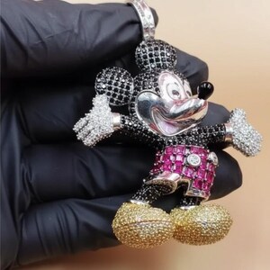 Puede incluir: Un colgante de Mickey Mouse con cara plateada, orejas y cuerpo con gemas negras, y manos con gemas blancas. Los pantalones cortos son rosas con gemas rojas y los zapatos dorados. El colgante lo sostiene una mano enguantada de negro.