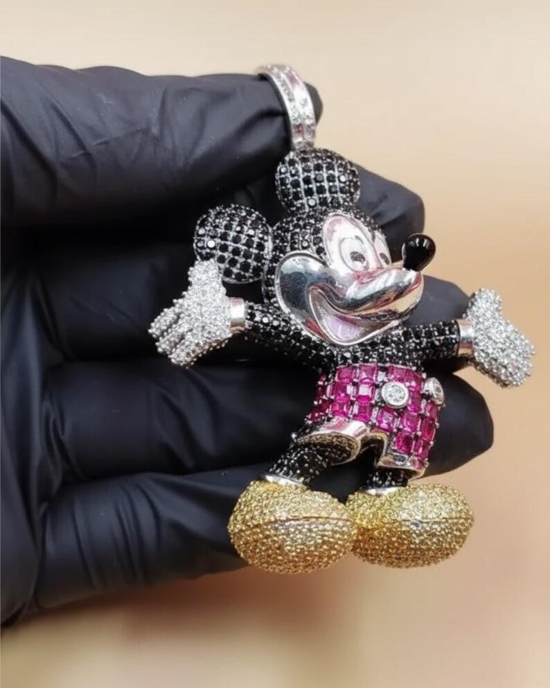 Puede incluir: Un colgante de Mickey Mouse con cara plateada, orejas y cuerpo con gemas negras, y manos con gemas blancas. Los pantalones cortos son rosas con gemas rojas y los zapatos dorados. El colgante lo sostiene una mano enguantada de negro.