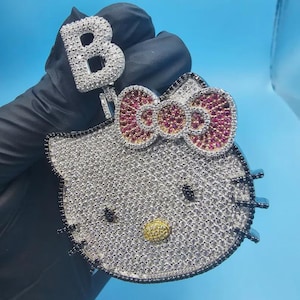 Puede incluir: Un colgante plateado con forma de gato de dibujos animados con un lazo, incrustado con pequeñas piedras brillantes. El lazo es rojo y la nariz del gato es dorada. Se adjunta un colgante separado con la letra "B".