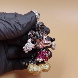 Puede incluir: Un colgante de Mickey Mouse con una fianza plateada. La figura est&aacute; adornada con piedras preciosas negras, blancas, rojas y doradas. El colgante presenta una cabeza negra, un atuendo rojo y negro y zapatos dorados. Las manos y las orejas son blancas.