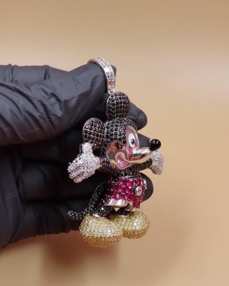 Puede incluir: Un colgante de Mickey Mouse con una fianza plateada. La figura est&aacute; adornada con piedras preciosas negras, blancas, rojas y doradas. El colgante presenta una cabeza negra, un atuendo rojo y negro y zapatos dorados. Las manos y las orejas son blancas.