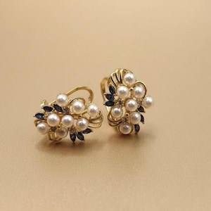 Op de afbeelding: Een paar goudkleurige oorbellen met clusters van witte parels en kleine, donkerblauwe edelstenen. De oorbellen hebben een bloemenontwerp, waarbij de parels zo zijn gerangschikt dat ze op bloemknoppen lijken. De achtergrond is lichtbruin.