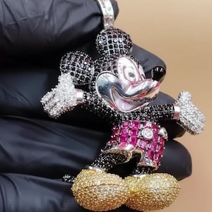 Puede incluir: Colgante de Mickey Mouse con cara plateada, detalles en blanco y negro y detalles en rojo y dorado. El colgante est&aacute; incrustado con piedras preciosas y tiene una argolla para una cadena.