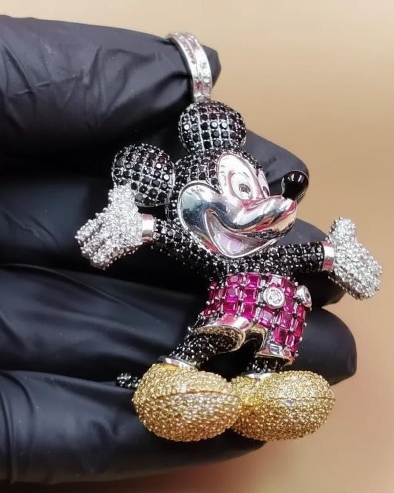 Puede incluir: Colgante de Mickey Mouse con cara plateada, detalles en blanco y negro y detalles en rojo y dorado. El colgante est&aacute; incrustado con piedras preciosas y tiene una argolla para una cadena.
