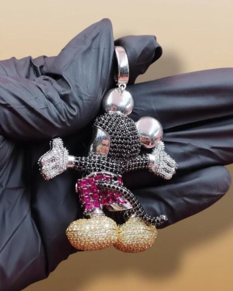Puede incluir: Un colgante de Mickey Mouse con una fianza plateada. El colgante presenta una cabeza negra y plateada, brazos con pulgares hacia arriba y pies dorados. Los pantalones cortos son de color magenta. El colgante est&aacute; sujeto por una mano enguantada.
