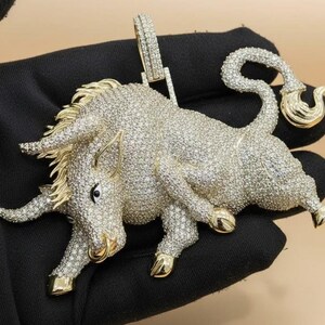 Könnte beinhalten: Ein diamantbesetzter Bullenanhänger mit goldenen Akzenten. Der Bulle wird in einer dynamischen Pose dargestellt, mit gekräuseltem Schwanz und nach vorne gerichteten Hörnern. Der Anhänger befindet sich vor einem schwarzen Hintergrund.