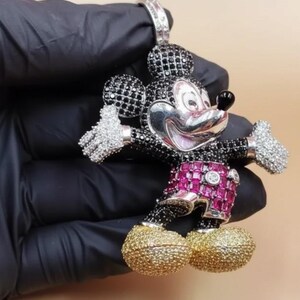 Puede incluir: Un colgante de Mickey Mouse con una cara plateada, detalles en blanco y negro y detalles en rosa y dorado. El colgante est&aacute; incrustado con piedras preciosas y tiene una argolla para una cadena.