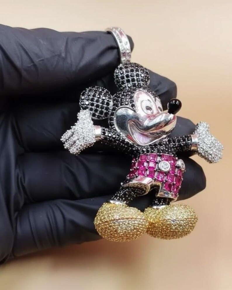 Puede incluir: Un colgante de Mickey Mouse con una cara plateada, detalles en blanco y negro y detalles en rosa y dorado. El colgante est&aacute; incrustado con piedras preciosas y tiene una argolla para una cadena.