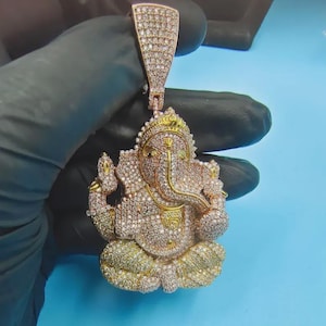 Può includere: Un ciondolo dorato raffigurante la divinità induista Ganesha, incastonato con numerose piccole pietre scintillanti. Il ciondolo presenta un design dettagliato, con un'asola nella parte superiore per una catena. La combinazione di colori è dorata, con accenti bianchi e gialli.
