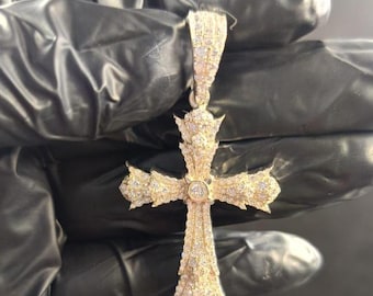 May include: A gold-colored pendant in the shape of a cross, encrusted with numerous small, sparkling gemstones. The cross has ornate detailing and a loop at the top for a chain. The pendant is held against a dark background.