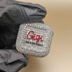 May include: A square, silver accessory covered in small, sparkling stones. The word "Gigi" is written in red cursive script across the centre, with "DA DON DADA" below. The accessory is held against a dark background.