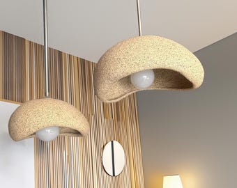 May include: Two pendant lights featuring textured, beige, dome-shaped shades with an irregular, organic edge. Each shade is suspended by a thin, silver-toned metal rod and contains a single, round, white light bulb centred within the opening.