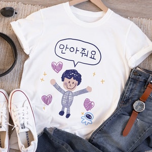 May include: White t-shirt with a cartoon illustration of a person with arms outstretched, surrounded by hearts and the Korean text "안아줘요" in a speech bubble. The shirt is paired with blue jeans, white sneakers, and a watch.