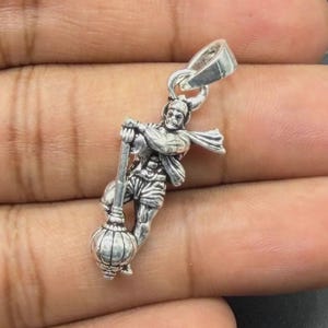 May include: A silver-toned metal pendant featuring a detailed figure of a deity holding a large mace. The figure is depicted in a dynamic stance with a flowing garment and a prominent bail at the top for attachment to a chain or cord.